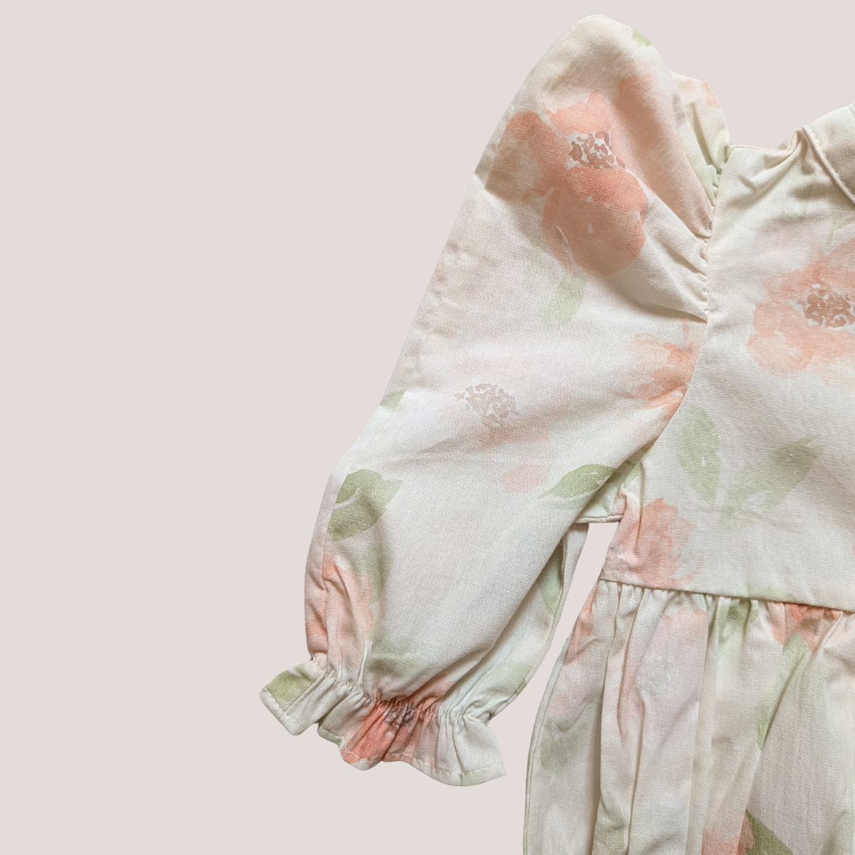 A close-up of a cotton baby romper featuring a soft, pastel floral print with dusty pink flowers and green leaves on a warm cream background. The romper has long, gathered puff sleeves with ruffled cuffs and a ruffled waist seam.
