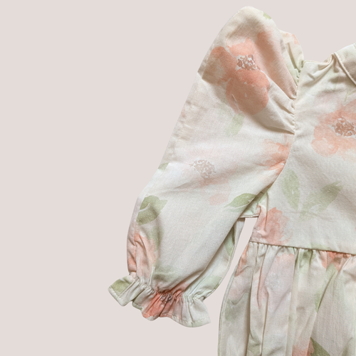 A close-up of a cotton baby romper featuring a soft, pastel floral print with dusty pink flowers and green leaves on a warm cream background. The romper has long, gathered puff sleeves with ruffled cuffs and a ruffled waist seam.