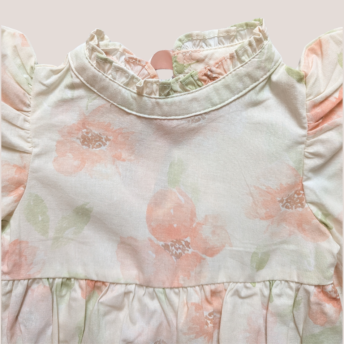 A close-up of a cotton baby romper featuring a soft, pale pink and sage green floral print on a cream-colored fabric. The romper has a ruffled mock neck, ruffled sleeves, and a ruffled waistband.