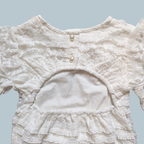 Close-up of the back of a white lace baby bodysuit, a round neckline, two decorative buttons, and a ruffled cutout at the waist.