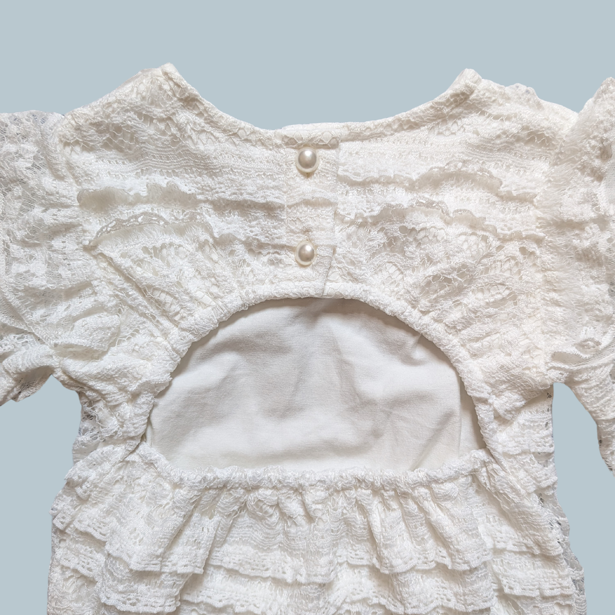 Close-up of the back of a white lace baby bodysuit, a round neckline, two decorative buttons, and a ruffled cutout at the waist.