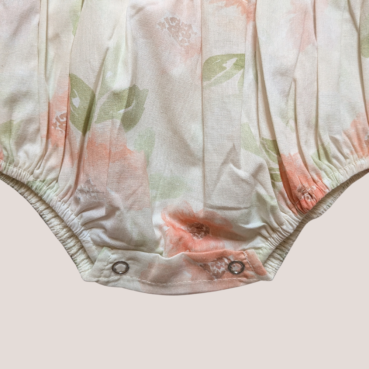 Close-up of a baby romper or bodysuit bottom with a soft floral print in peach, sage green, and cream, finished with snap closures at the leg openings.
