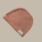 A terracotta ribbed knit baby beanie hat with a small white fabric brand tag for Ela & Ren