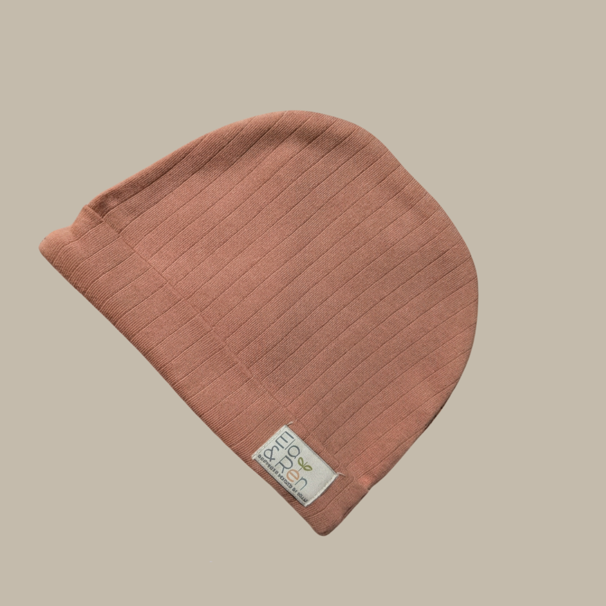 A terracotta ribbed knit baby beanie hat with a small white fabric brand tag for Ela & Ren