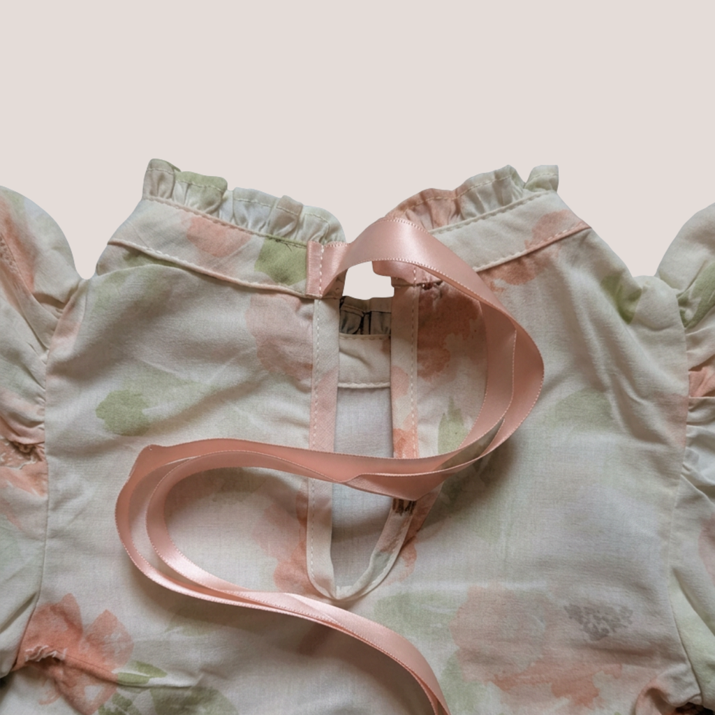 A close-up of a baby romper with ruffled shoulders, a pale pink and sage green floral print, and a light pink satin ribbon tied around the neckline.