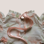 A close-up of a baby romper with ruffled shoulders, a pale pink and sage green floral print, and a light pink satin ribbon tied around the neckline.